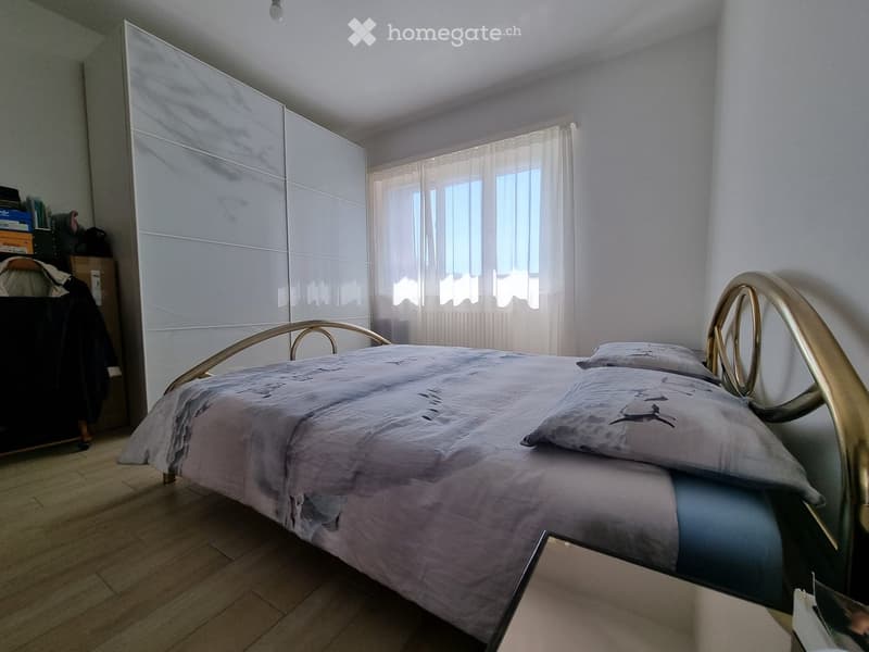 Bedroom with a bed, two pillows, white walls, wooden floor, white curtains, white wardrobe, and a window.