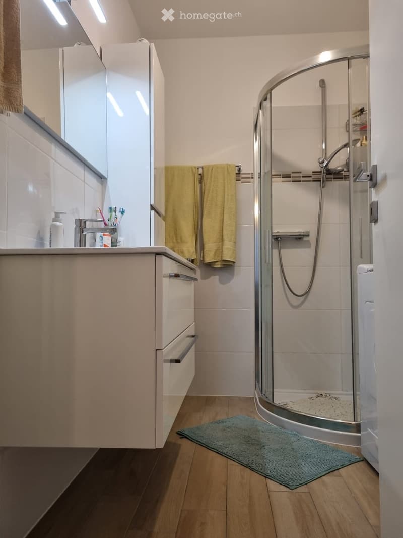 modern bathroom with white tiles, glass shower, white vanity with drawers, yellow towels, green rug, wooden floor