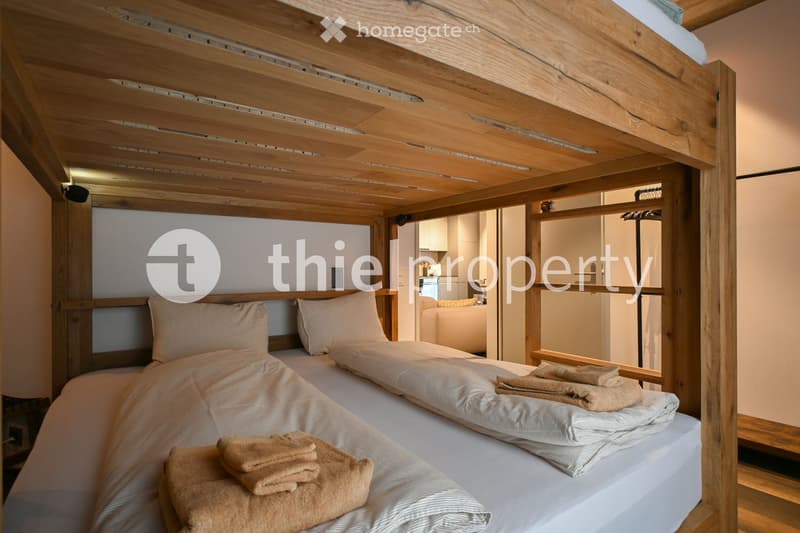 Double bunk bed with beige linen, white bedsheets, and beige throw pillows. Two stacks of towels on the lower bed. Glass window at the back. Wooden ceiling and walls.