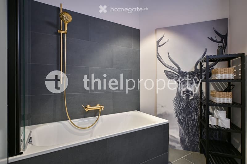 modern bathroom with white bathtub, shower, gold fixtures, black tiled wall, wallpaper with deer image, shelving unit with basket and toilet paper