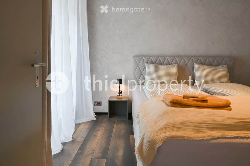 Bedroom: grey walls, wooden floor, twin beds, nightstand, decorative lamps, curtains, gray headboards, plaid cushions, towels