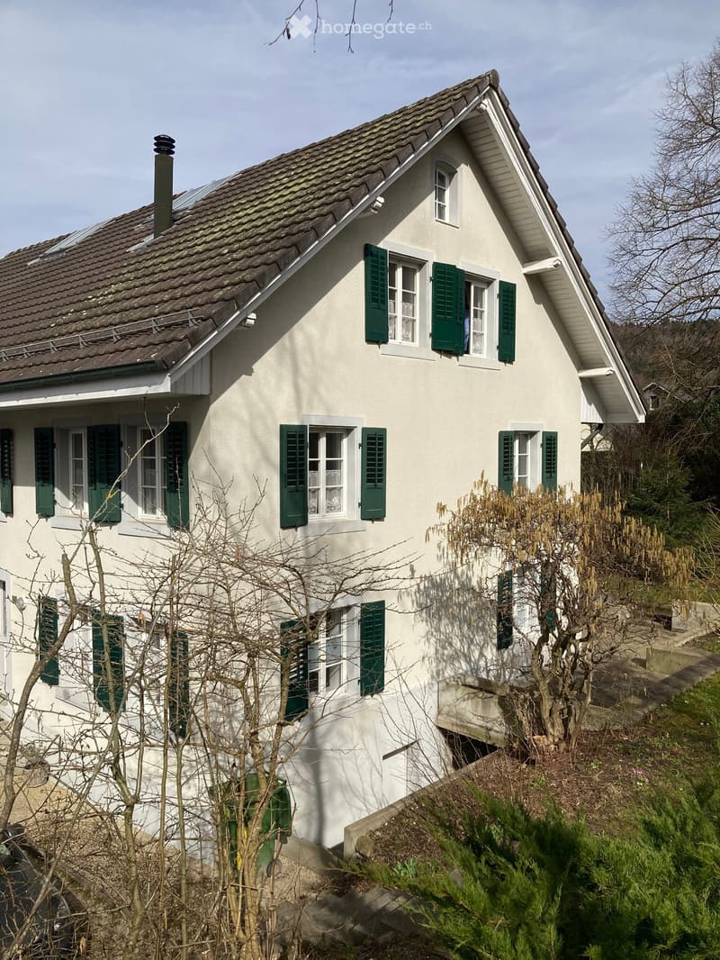 Two-story house with white walls, green shutters, and a tiled roof. The house has multiple windows and a chimney. There is a small garden area in front with some bare trees and shrubs.