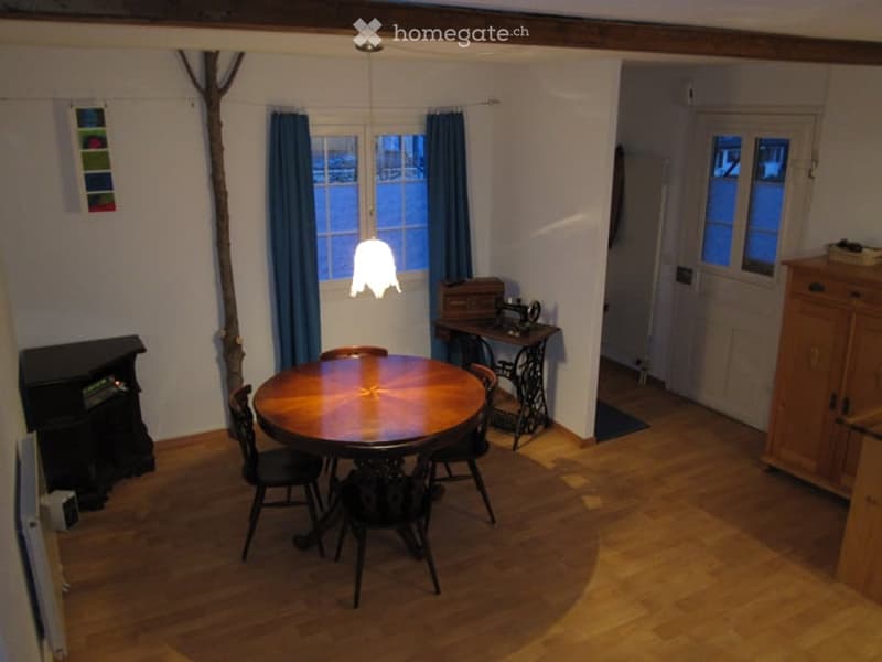 Cozy dining area with a round wooden table, four chairs, a hanging pendant light, a sewing machine on a side table, and a small fireplace. The room has wooden flooring and large windows with blue curtains.