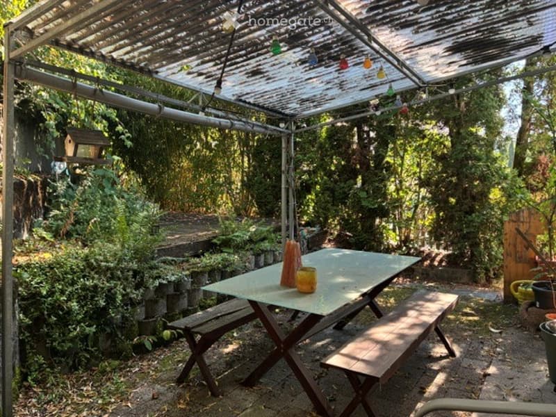 A covered outdoor seating area with a picnic table, benches, and hanging string lights, surrounded by lush greenery and plants.