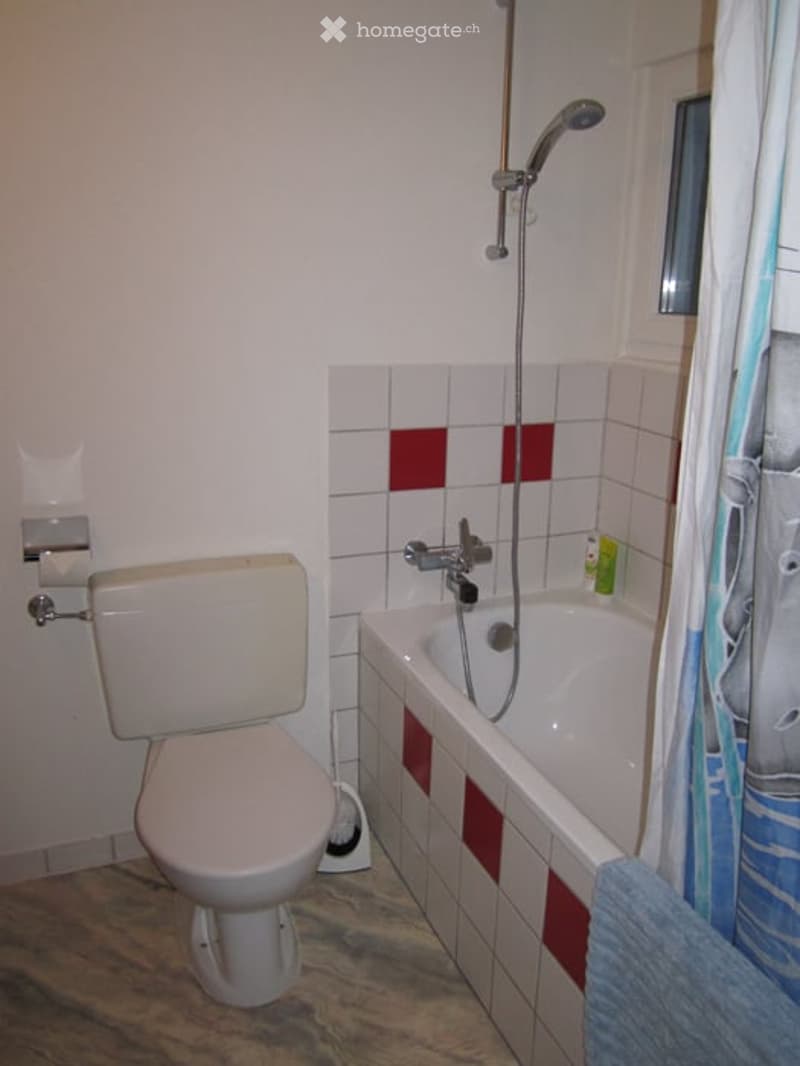 Bathroom with a toilet, bathtub, and showerhead. The walls are tiled with white and red accents, and a small window is visible on the right side.