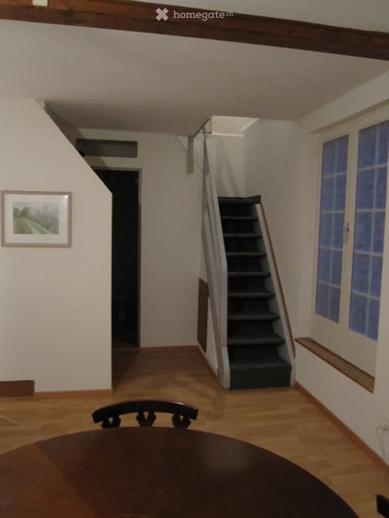 A small, well-lit room with wooden flooring, a staircase leading upwards, a chair, and a table visible in the foreground.