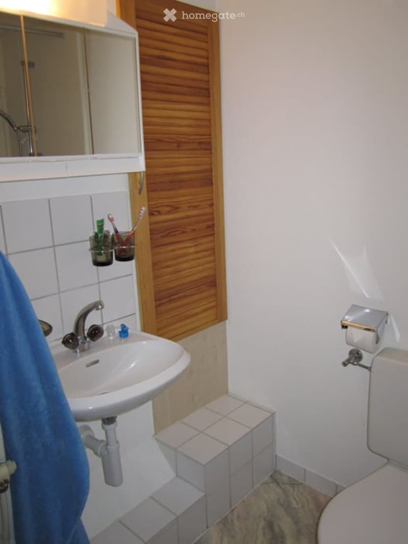A compact bathroom with a white sink, mirror, and toilet. The sink has a modern faucet, and toothbrush holders are mounted on the wall. A blue towel hangs on the side, and the floor is tiled.