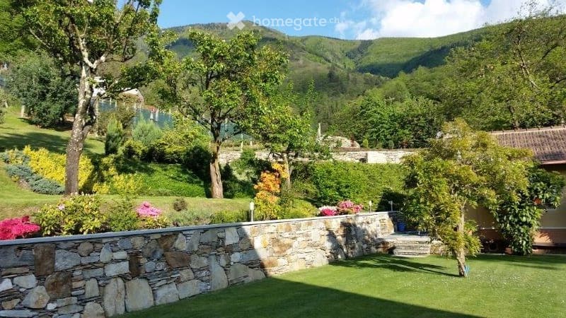 A well-maintained garden with a stone wall, green lawn, colorful flowers, and trees, overlooking a scenic mountainous landscape.