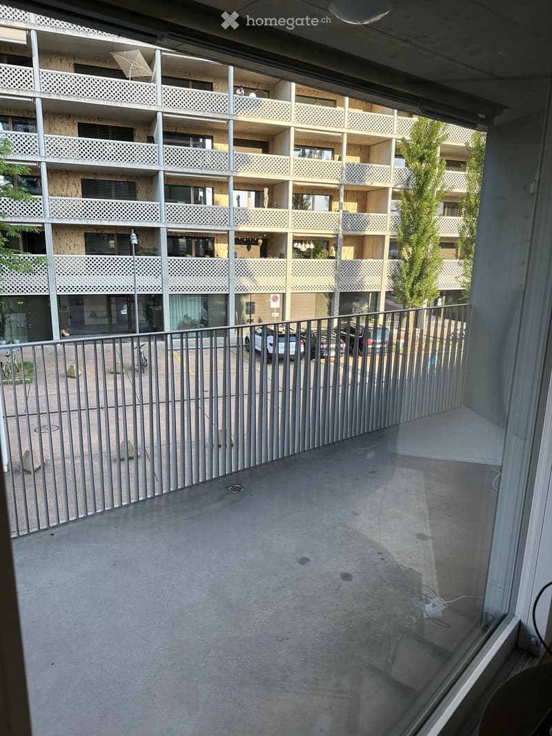 Balcony with metal railing overlooking a multi-story residential building with balconies and parked cars below.
