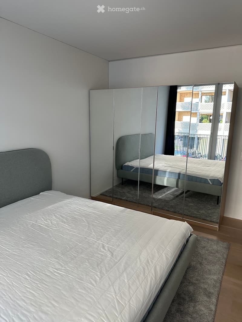 Modern bedroom with a large bed, white bedding, and a large mirrored wardrobe reflecting the bed and part of the room.