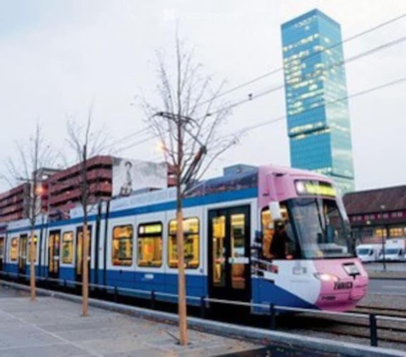 A modern tram with pink, blue, and white livery is parked on tracks in an urban setting, with a tall skyscraper visible in the background.