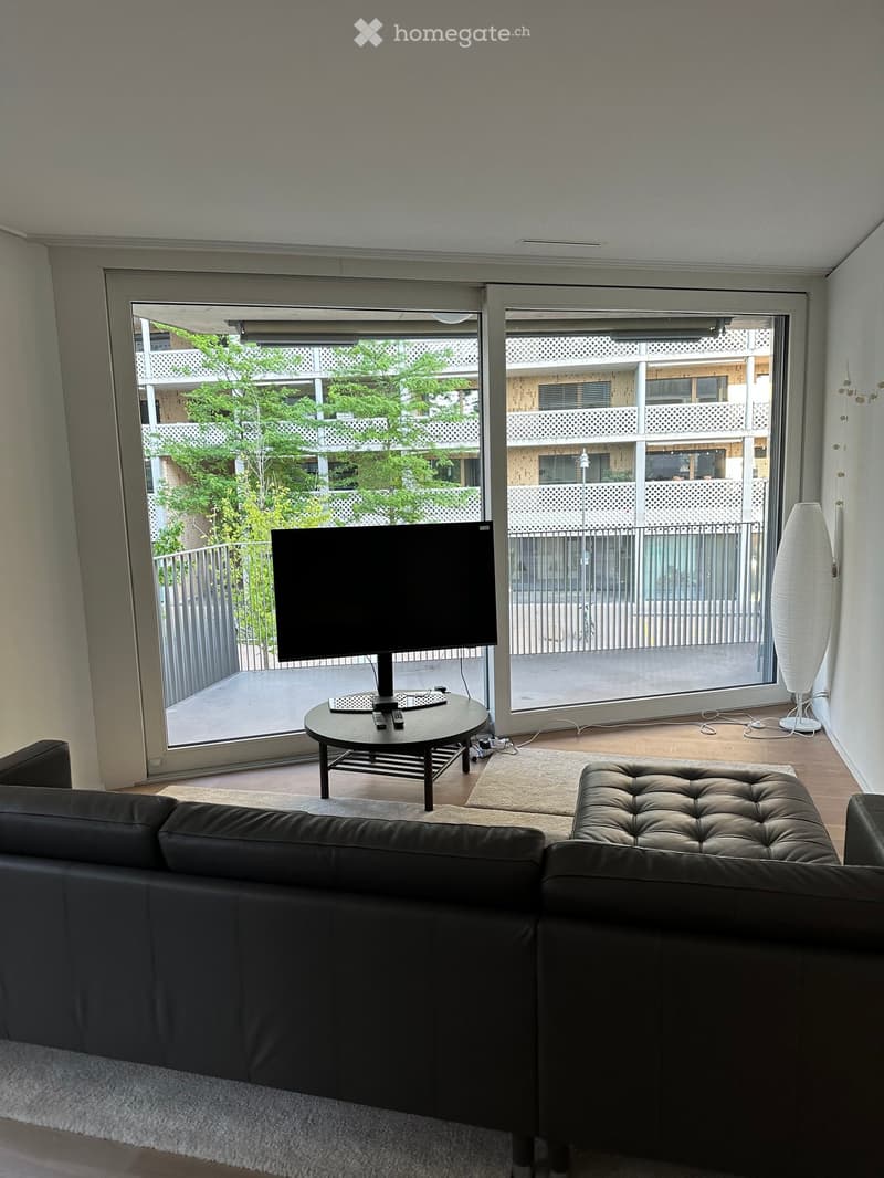 Modern living room with a black leather sofa, ottoman, coffee table, television, and floor lamp, featuring large sliding glass doors opening to a balcony.