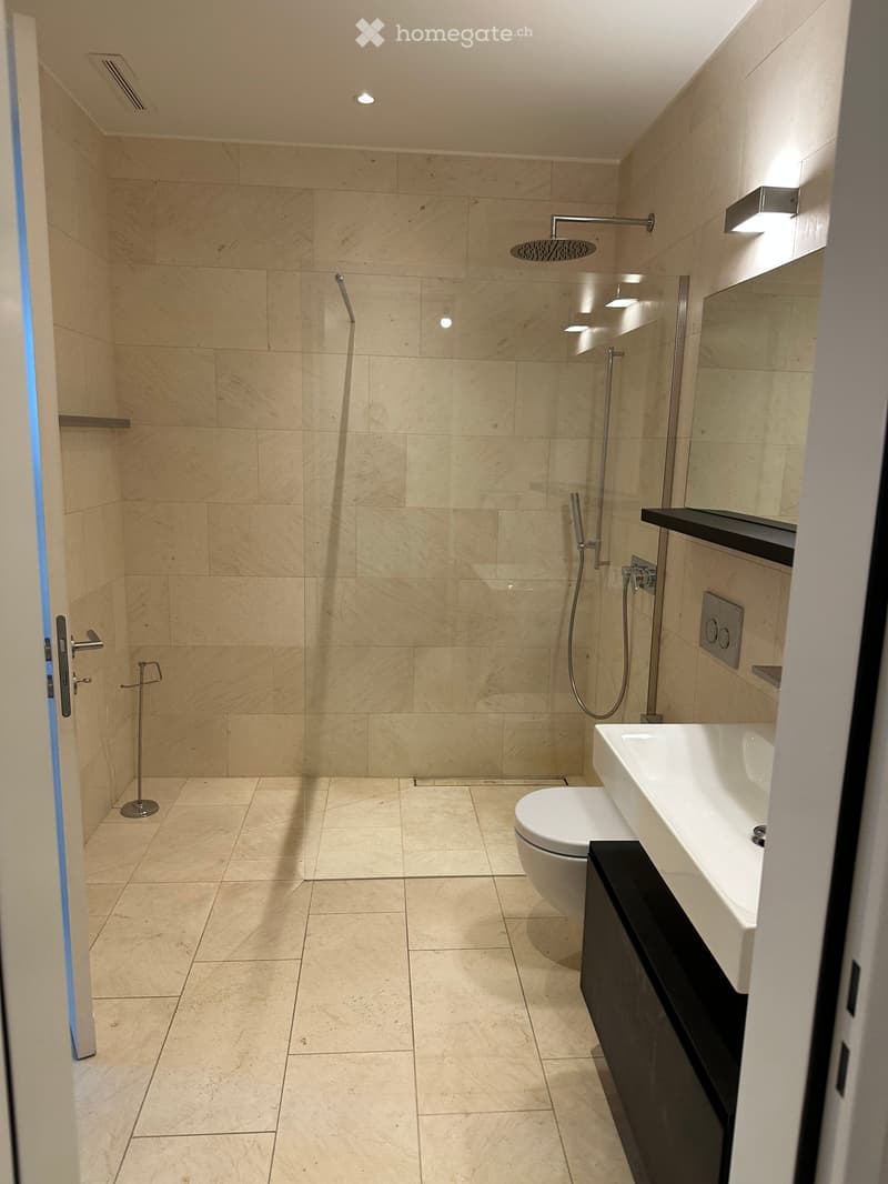 Modern bathroom with beige tiled walls and floor, featuring a shower area with glass enclosure, a toilet, a sink with a countertop, and a towel rack near the door.