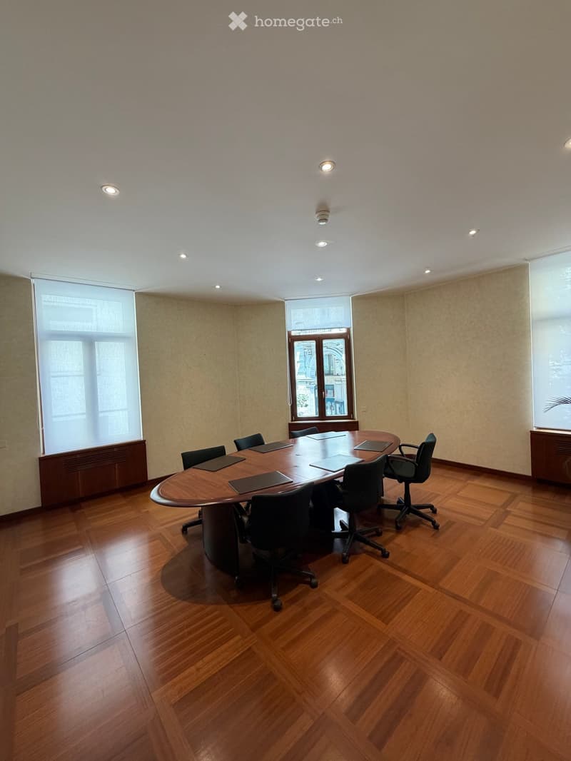 A conference room with a large wooden table and several black chairs arranged around it. The room features wooden flooring, large windows, and recessed lighting.