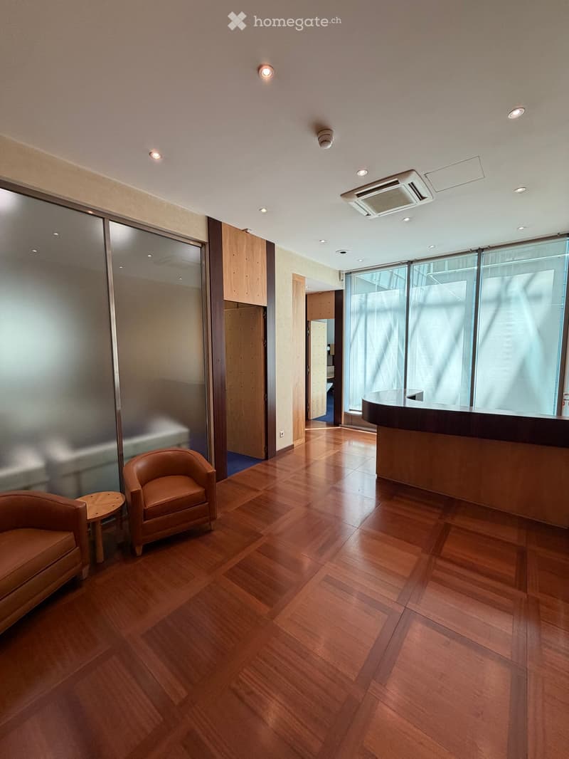 A modern, well-lit reception area with wooden flooring, a leather sofa, a small round table, a leather armchair, and a reception desk.