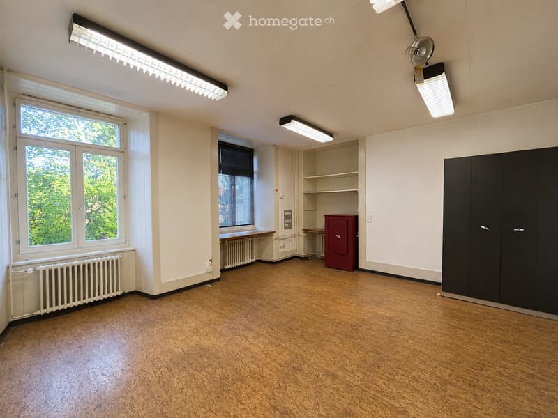 Empty room with wooden flooring, white walls, large windows, built-in shelving, and a small red cabinet.