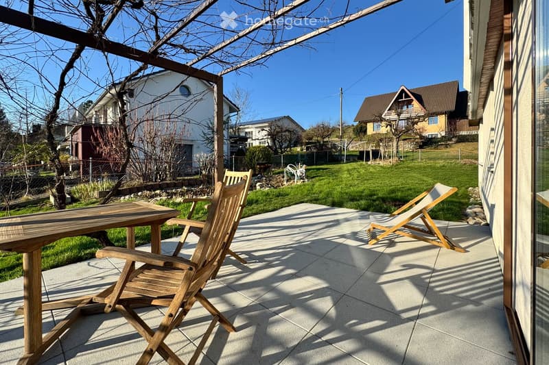 A sunny outdoor patio with a wooden table and two wooden chairs, surrounded by a grassy lawn and neighboring houses.