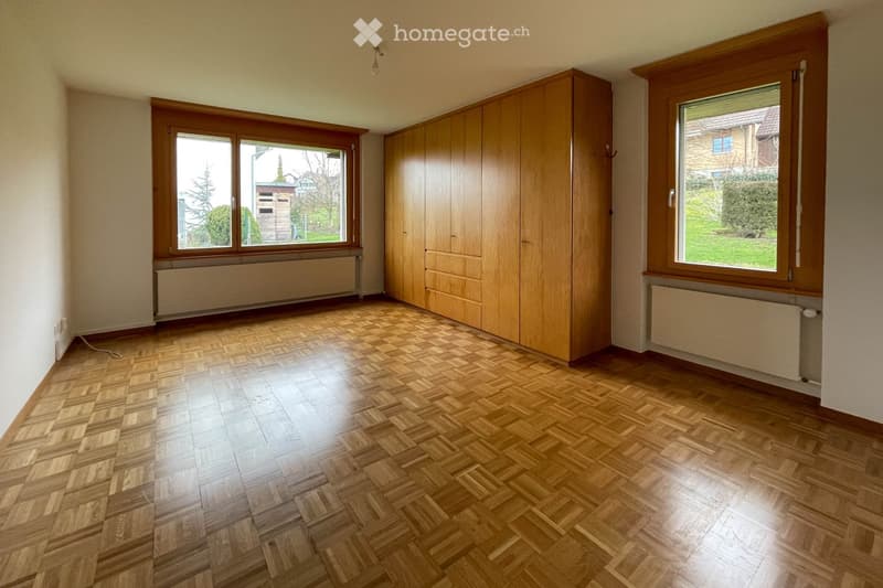 Empty room with wooden herringbone flooring, large windows with wooden frames, built-in wooden cabinetry, and radiators beneath the windows.
