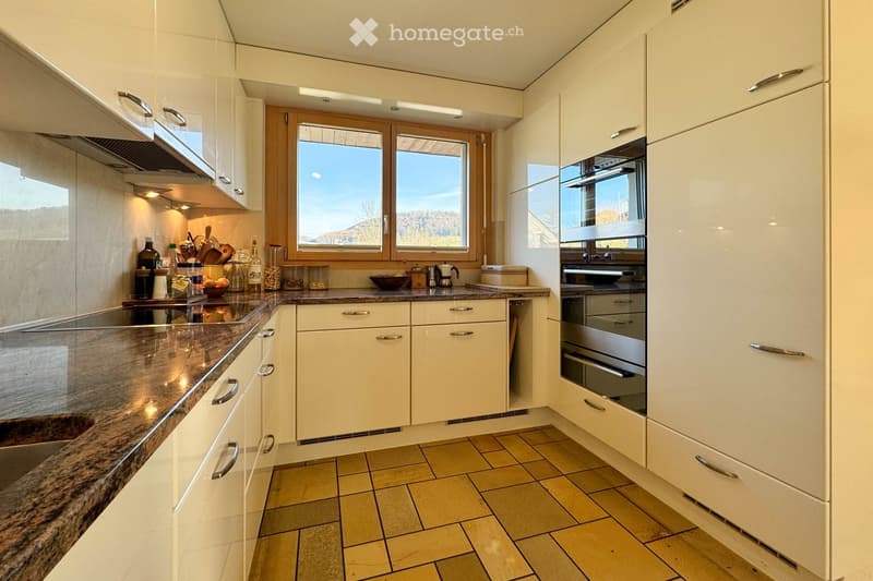 Modern kitchen with white cabinets, granite countertop, built-in oven, and various kitchen utensils on the counter. Large windows provide natural light and a scenic view.