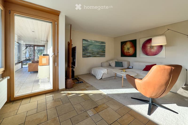 Modern living room with a grey sofa, coffee table, ottoman, and a leather chair. Large windows and a sliding glass door lead to an outdoor area. Artwork adorns the walls.