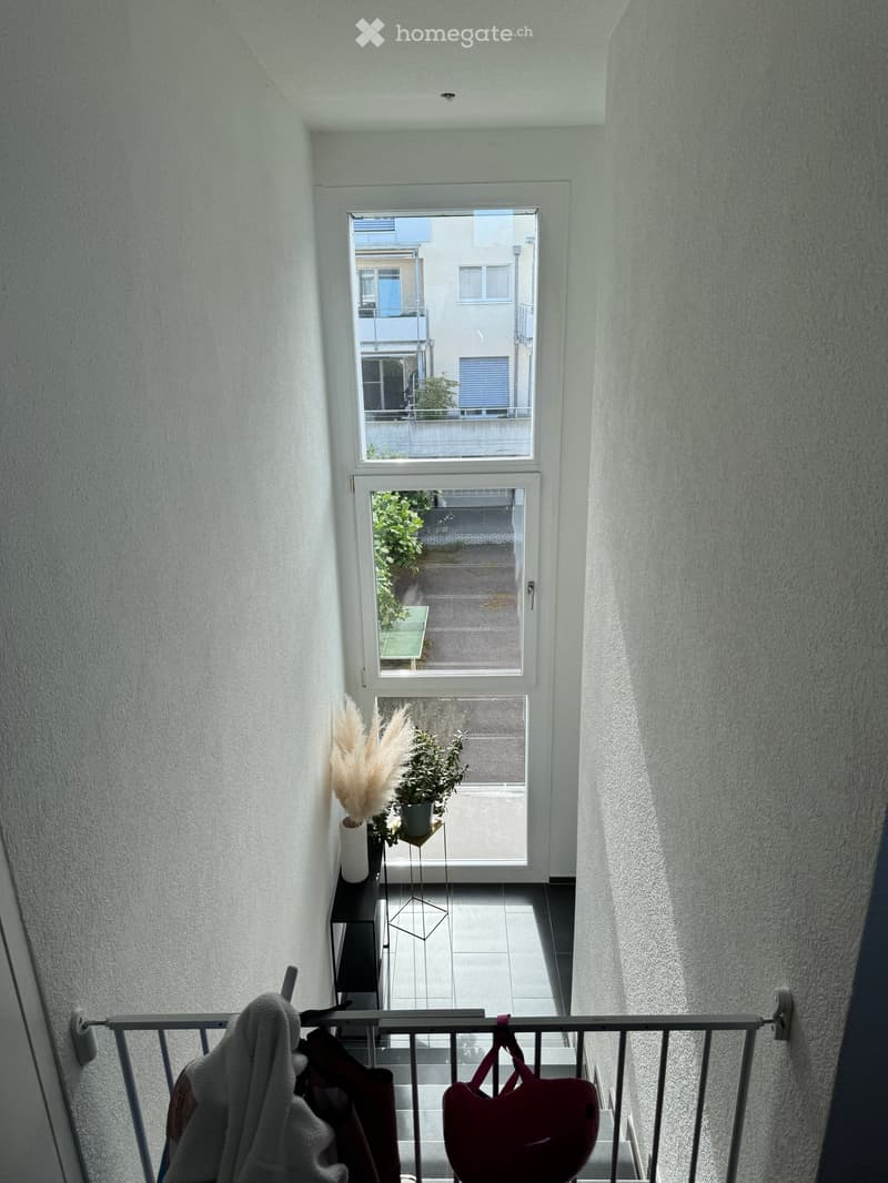 Balcony with a small table, potted plants, and a vase, overlooking a street with buildings and greenery.