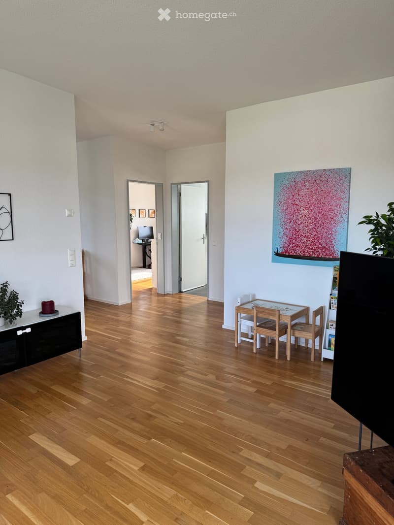 Bright, open living space with wooden flooring, a small dining table with chairs, a cabinet, a TV screen, and a colorful painting on the wall.