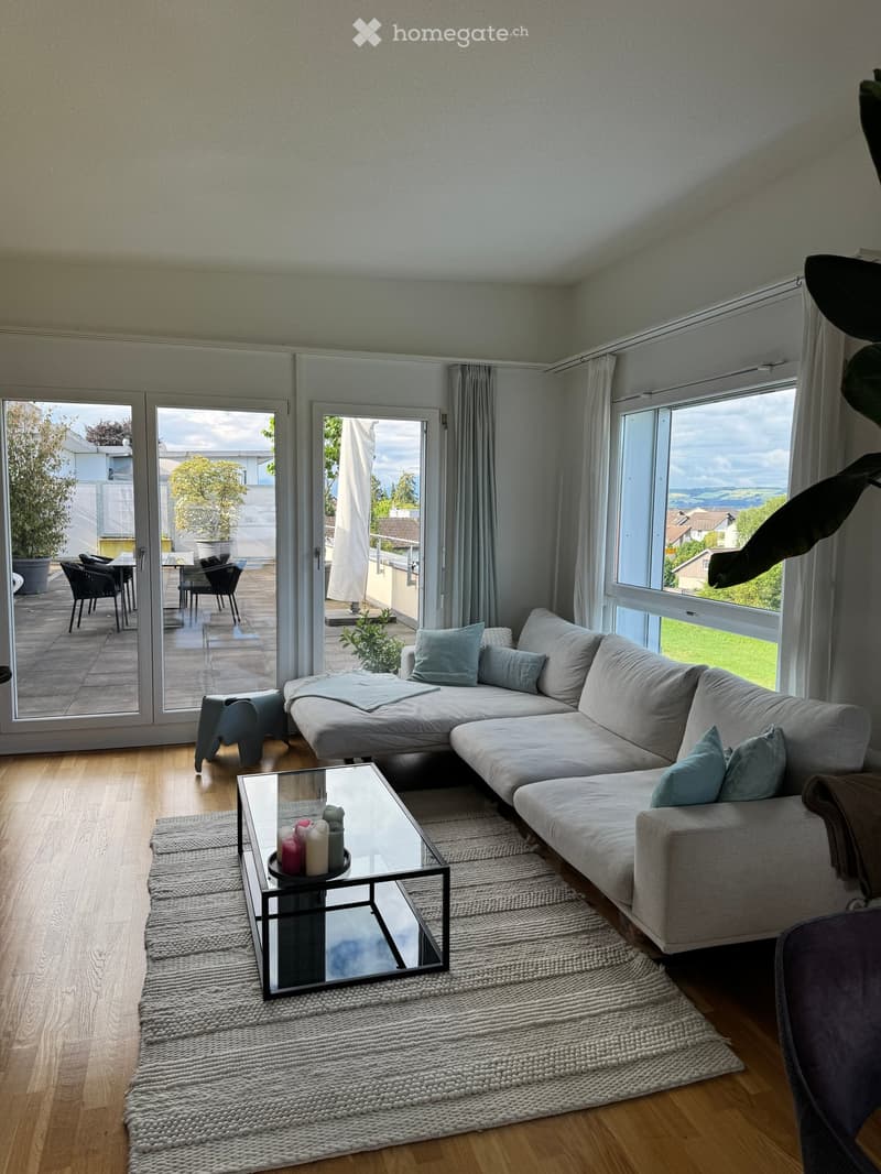 Modern living room with large windows and sliding glass doors leading to a balcony. Features a grey sectional sofa, glass coffee table with candles, a small stool, and a dining area with chairs and a table visible through the open doors.