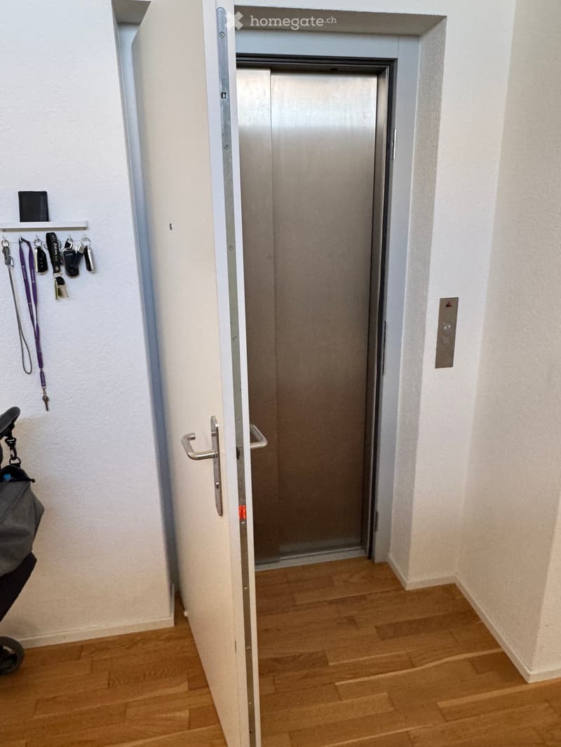 A hallway with wooden flooring, white walls, and an elevator door visible at the end. A small shelf with keys and a bag is on the left side.