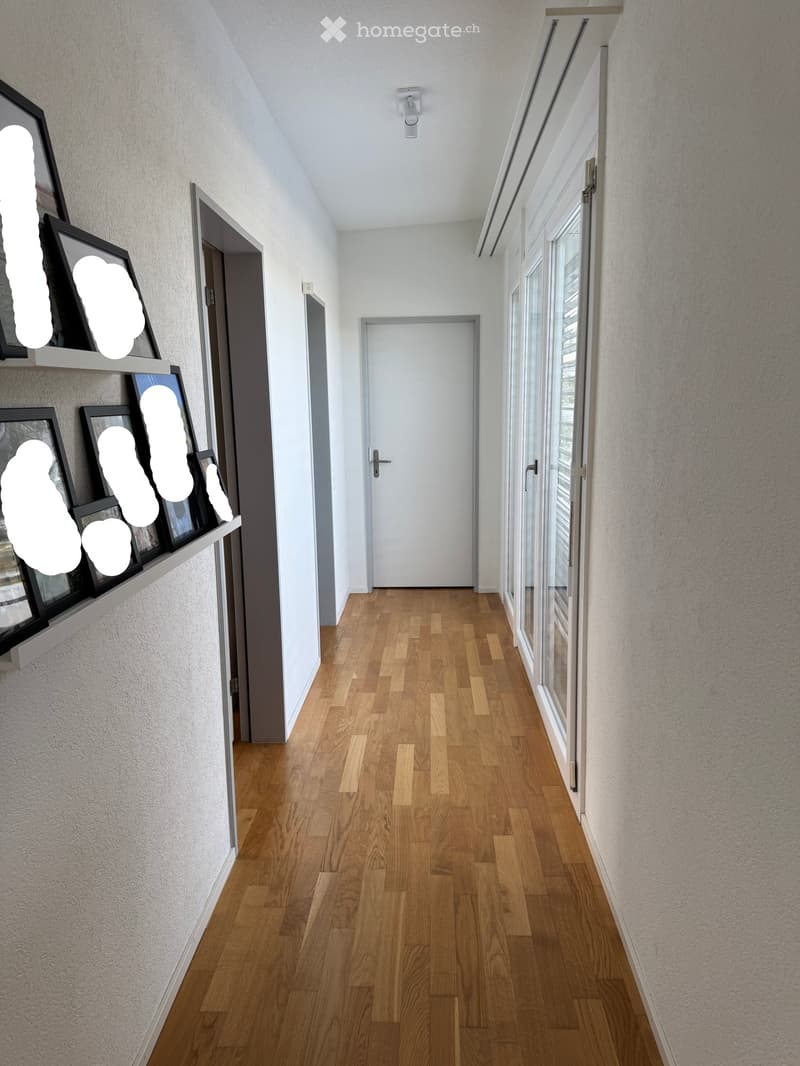 Modern hallway with wooden flooring, white walls, and large windows on one side. Features a white door at the end and a wall-mounted shelf with framed artwork on the left.
