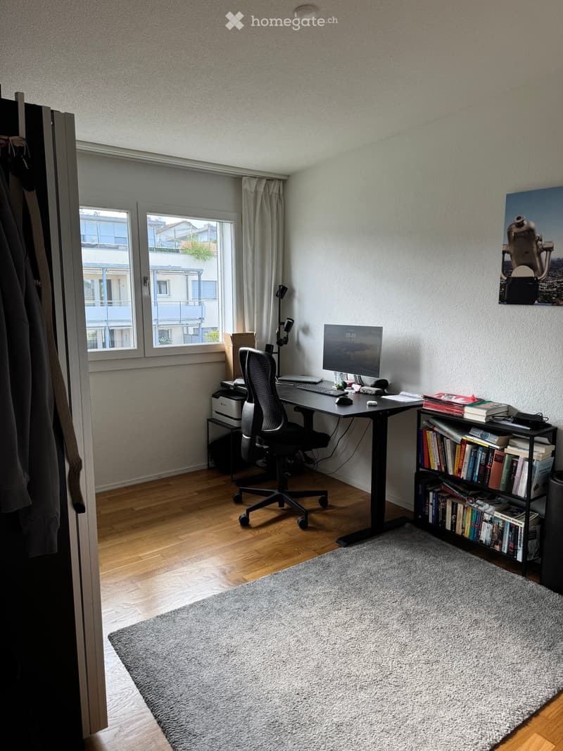 A modern, well-lit home office with a wooden floor, large window with curtains, desk with a monitor, keyboard, mouse, chair, bookshelf filled with books, and a grey rug.