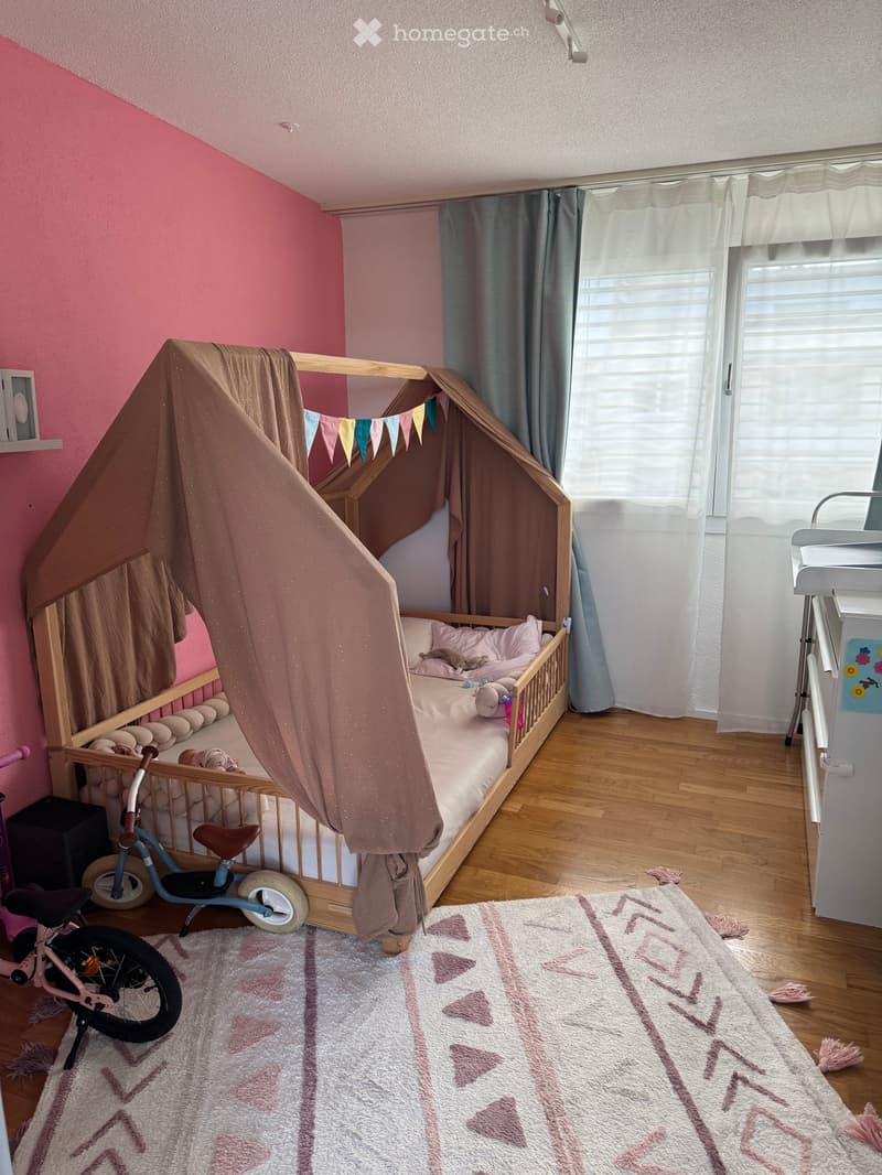 A cozy children's bedroom with a wooden bed, pink bedding, a curtained canopy, a small bicycle, and a decorative rug.