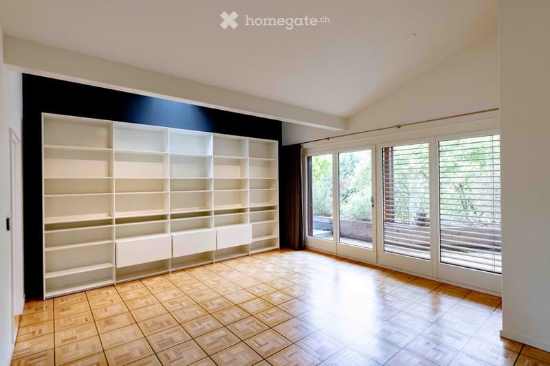 Empty room with wooden herringbone flooring, white built-in shelving unit, large windows with blinds, and a view of a garden area.