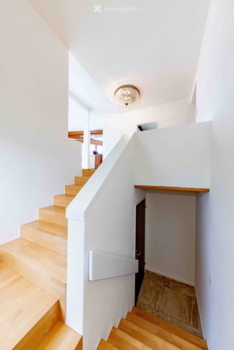 Modern staircase with wooden steps and white walls, featuring a chandelier and a small landing area with a shelf.