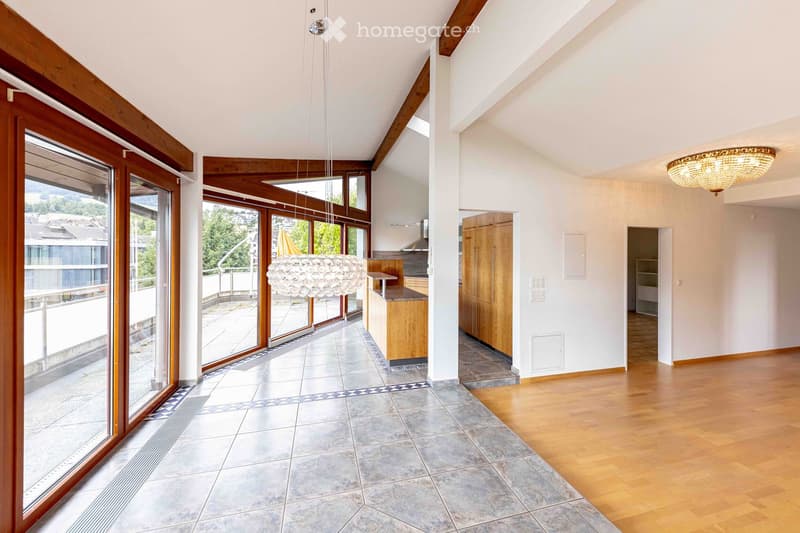 Bright, open hallway with wooden floors, large windows, and modern lighting fixtures. The space features a mix of tiled and wooden flooring, leading to a kitchen area with wooden cabinets and a countertop.
