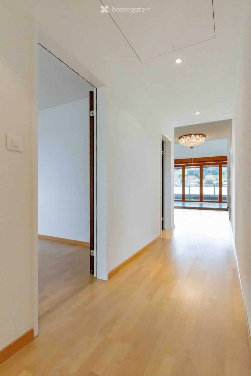 Empty hallway with light wooden flooring, white walls, and recessed ceiling lighting. Features a chandelier in the background and a view of a bright room beyond.