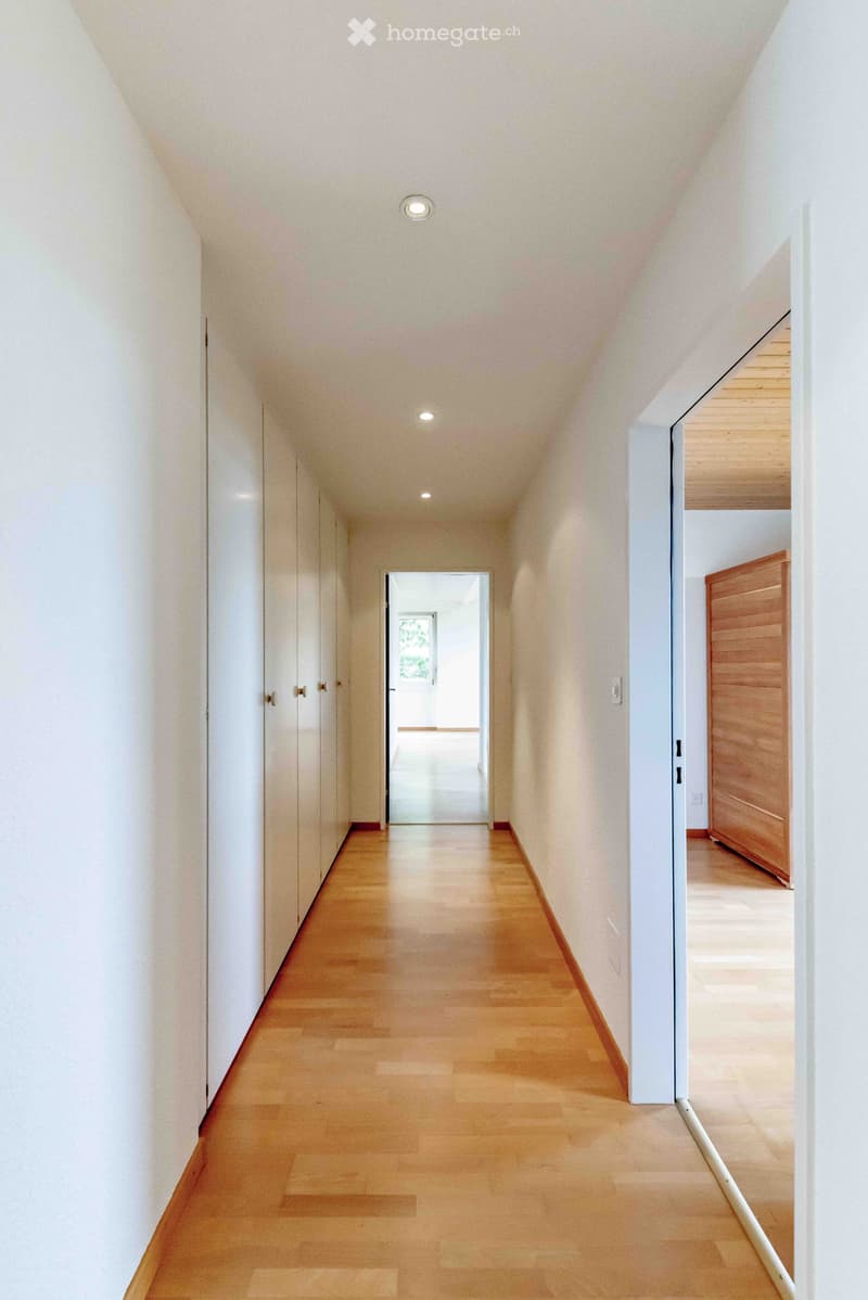 Modern hallway with light wooden flooring, white walls, and recessed ceiling lighting. Features a closed wooden door on the right and an open doorway leading to another room.