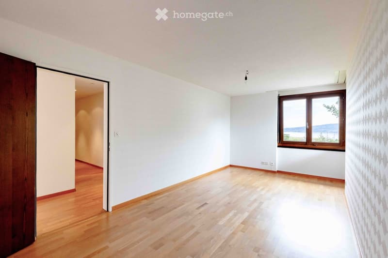 Empty room with wooden flooring, white walls, and a large window offering a scenic view. The room features a wooden door on the left and a light switch on the wall.