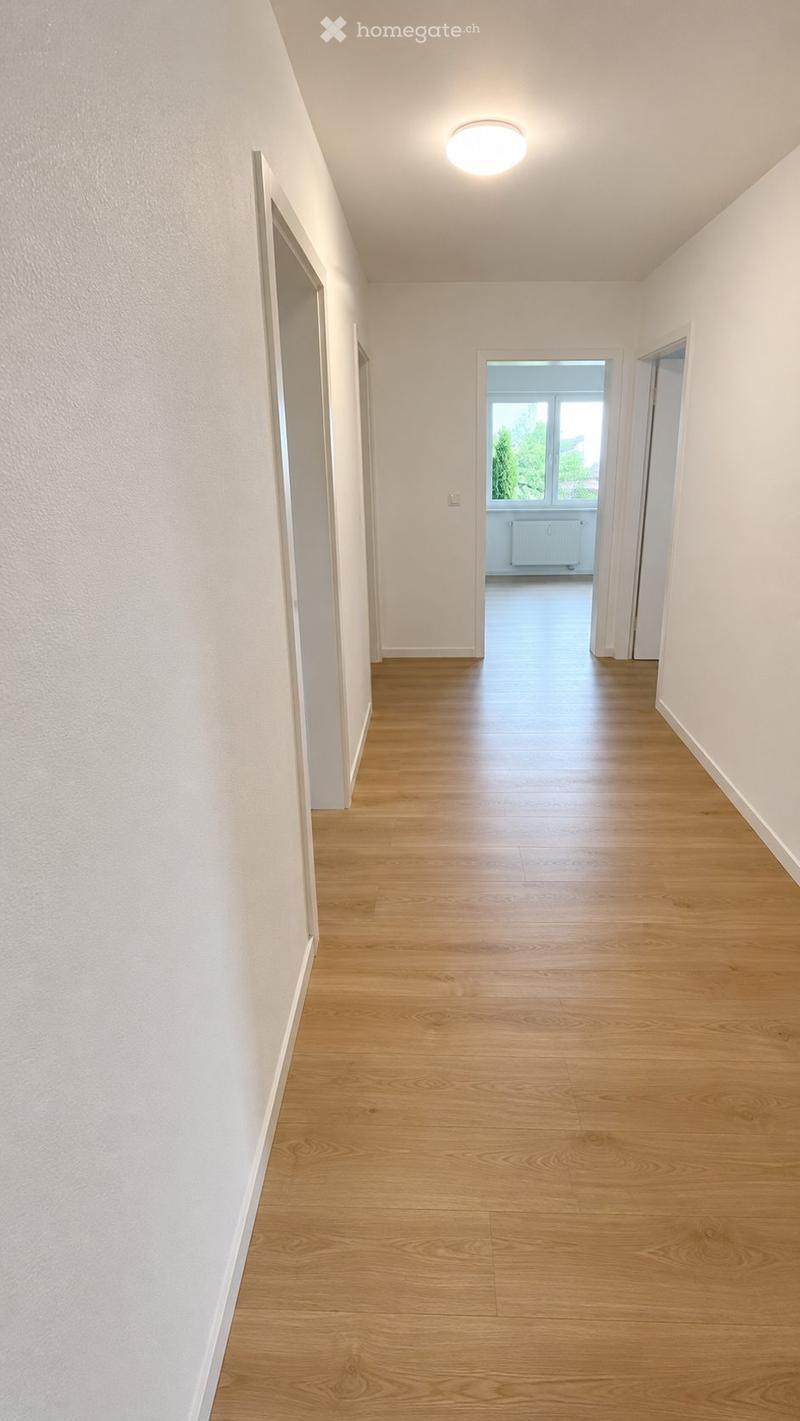 Empty hallway with light wooden flooring, white walls, and a ceiling light fixture. Two open doors lead to adjacent rooms, one with visible windows and a radiator.