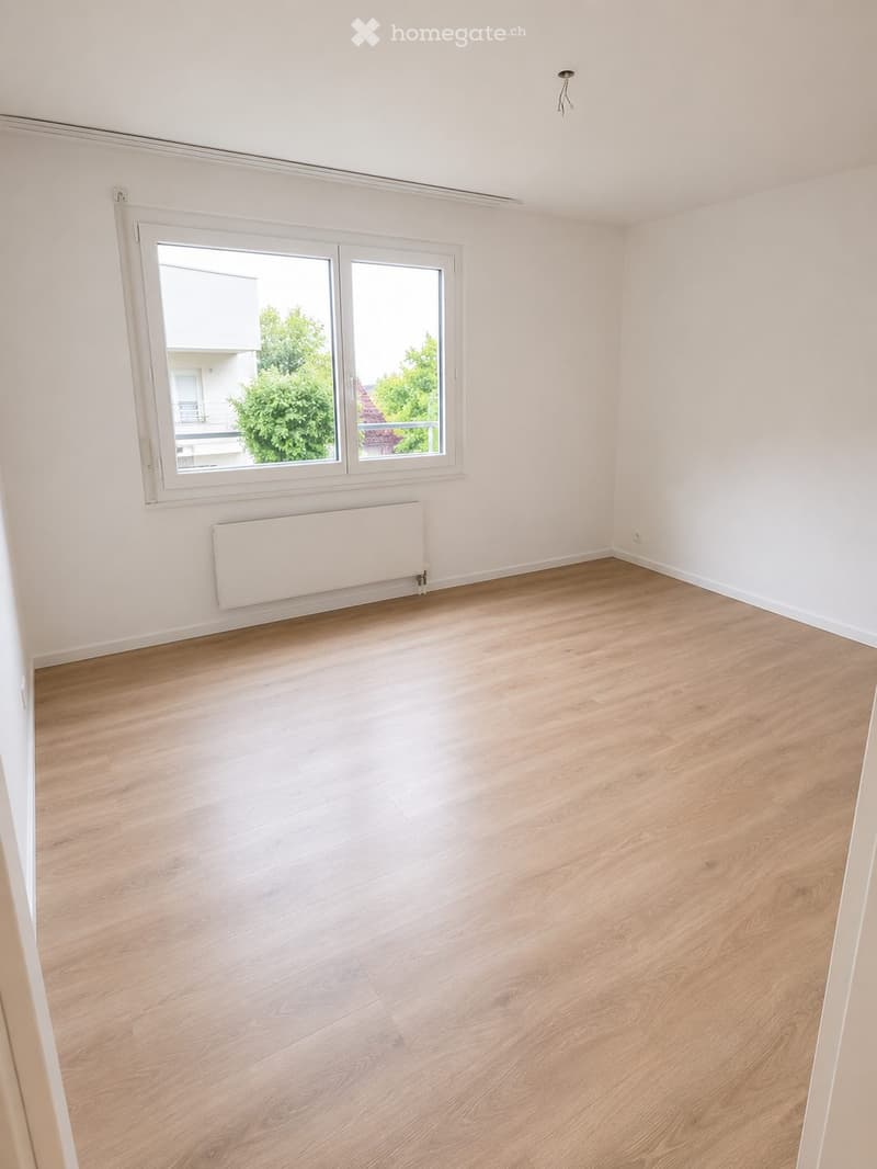Empty room with light wooden flooring, white walls, and a large window with a view of greenery outside. A radiator is mounted below the window.