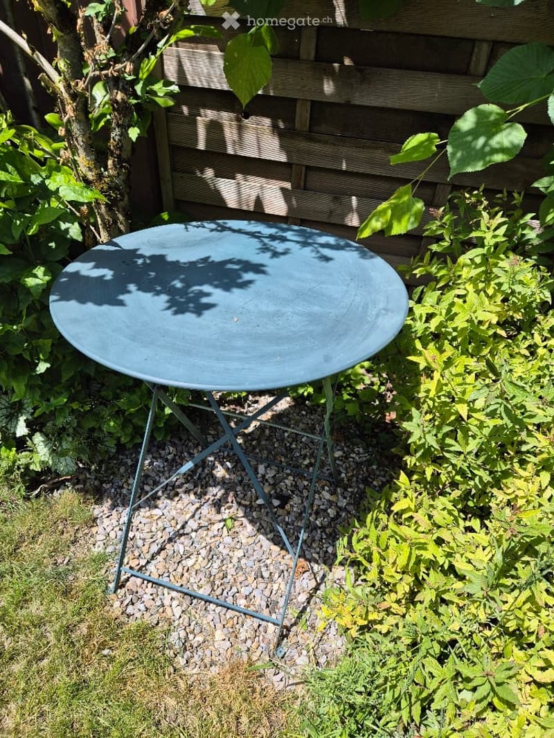 A small outdoor seating area with a round metal table and a tree, surrounded by greenery and a wooden fence.