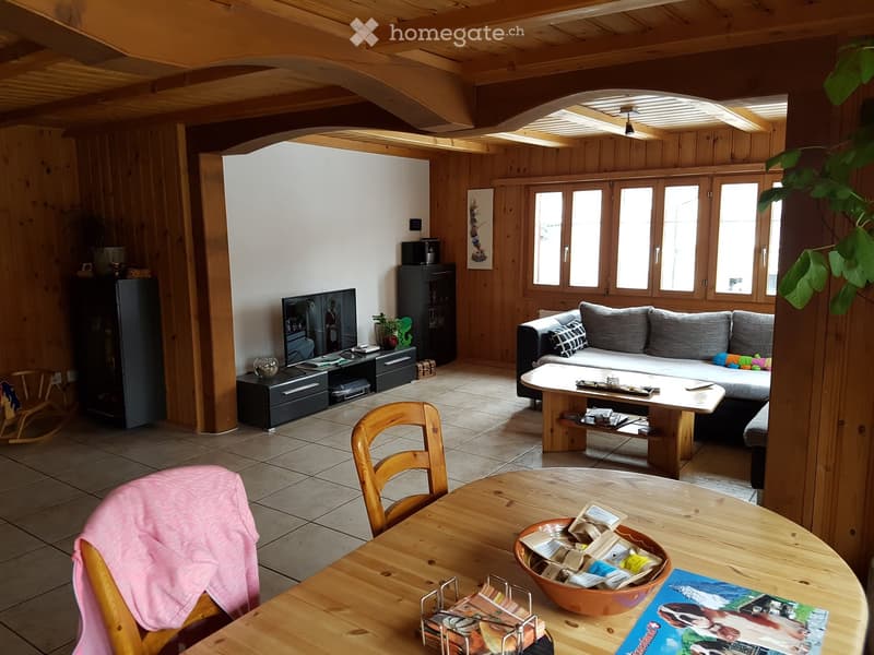 A cozy living room with wooden beams, a grey sofa, a wooden coffee table, a dining table with chairs, and a TV stand with a screen. The room features wooden walls, tiled flooring, and large windows letting in natural light.