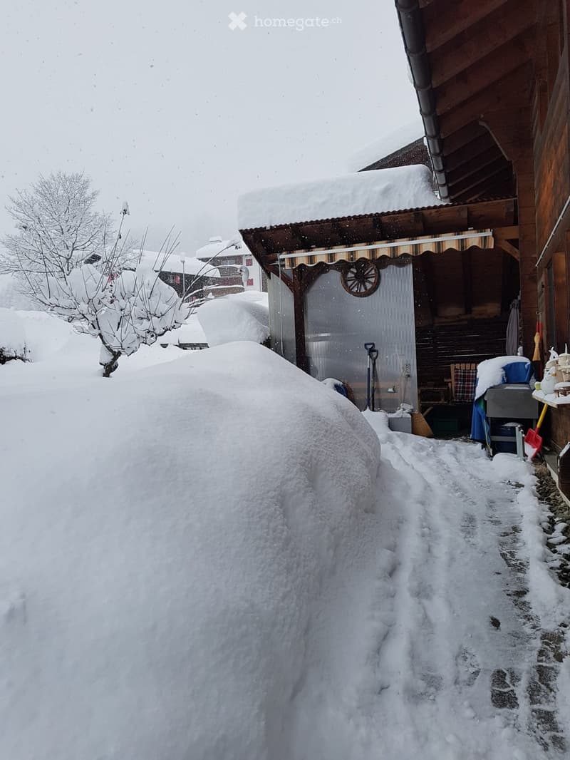 Snow-covered pathway leading to a wooden house with a covered entrance, surrounded by snow-covered trees and structures.