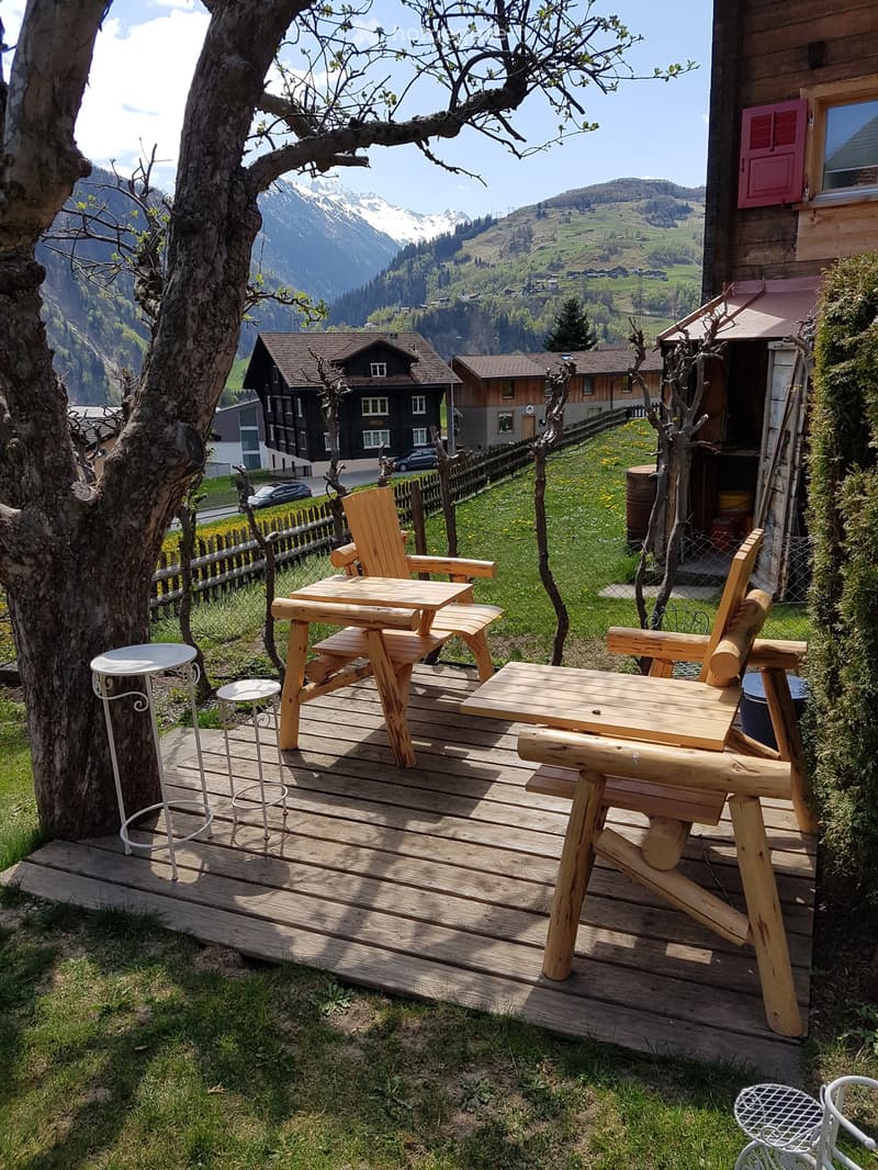 Wooden deck with a table, chairs, and stools, overlooking a scenic mountain valley with houses and a clear blue sky.