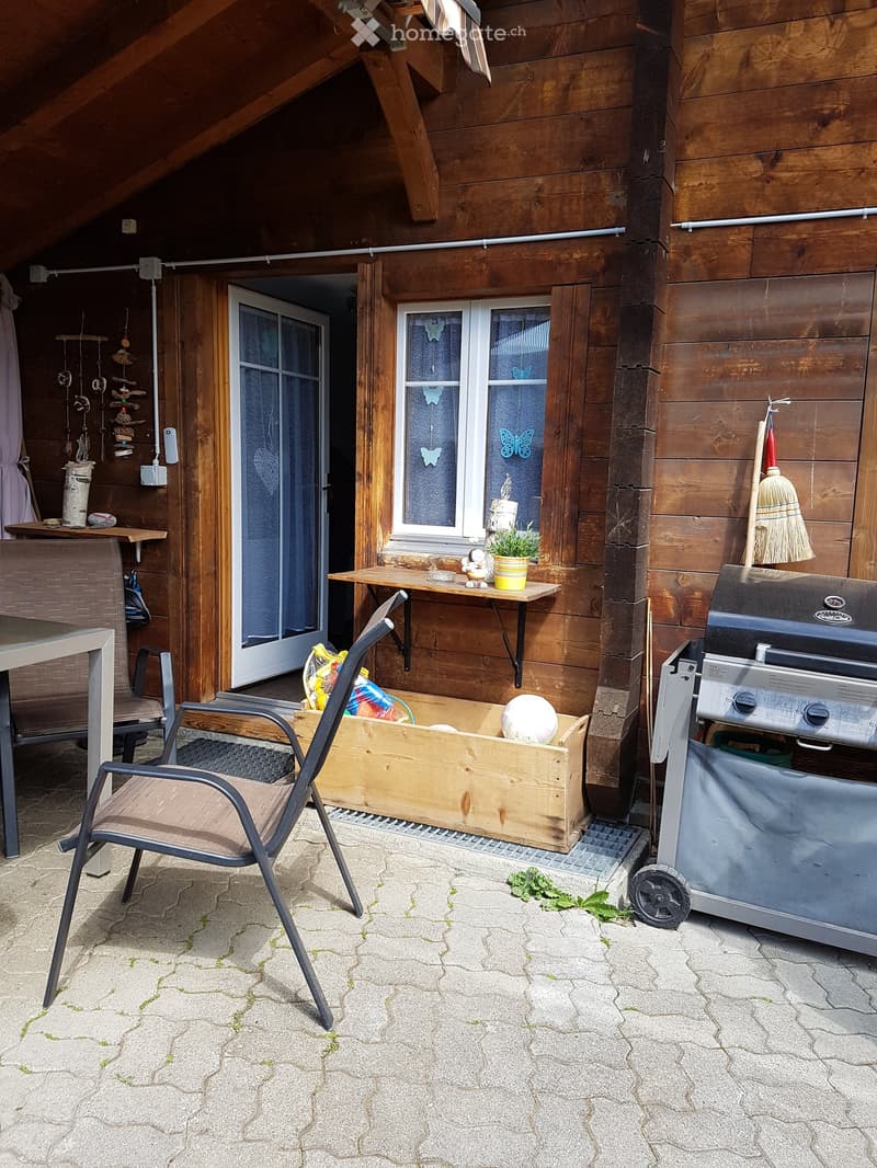 Wooden outdoor area with a slanted roof, featuring a chair, table, grill, wooden box, small plant, and a hanging curtain.