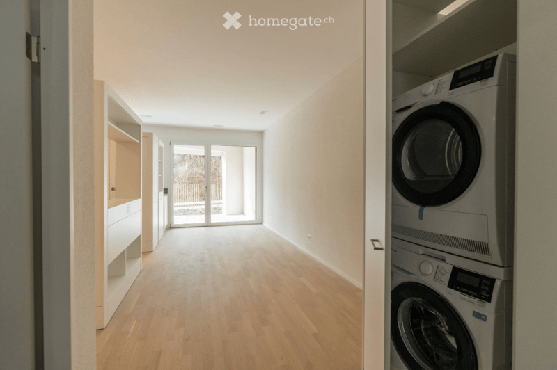 Empty hallway with wooden flooring, white walls, and a large glass door leading to an outdoor area. Laundry area visible with stacked washer and dryer.