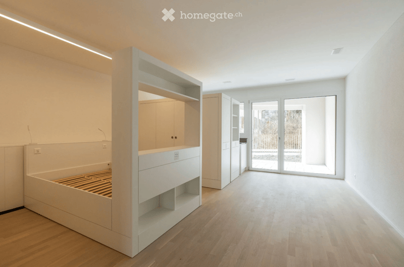 Empty room with light wooden flooring, white walls, built-in bed frame with shelves, and large glass doors leading to an outdoor area.