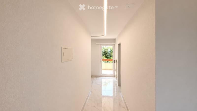 A clean, modern hallway with white walls, a polished marble floor, and a recessed ceiling light. A door at the end leads to a balcony or outdoor area.