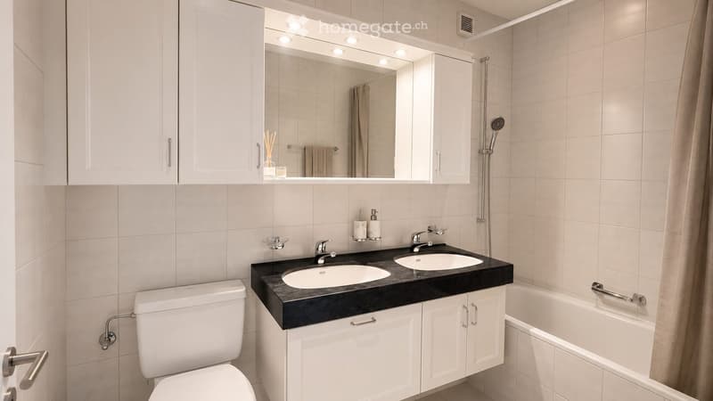 Modern bathroom with white tiles, double sink, toilet, bathtub, and a large mirror with built-in lighting.