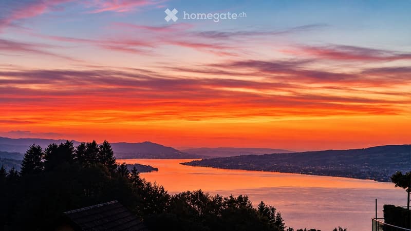 Panoramic view of a lake at sunset, with city lights visible across the water and distant mountains in the background.