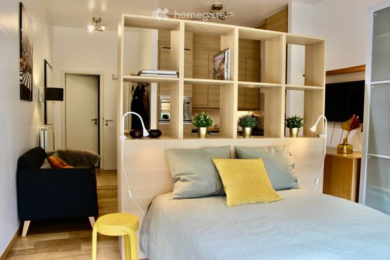Modern, open-plan living and sleeping area with a bed, sofa, shelves, and small potted plants. The space features a light-colored bed with multiple pillows, a small yellow stool, and a dark-colored sofa with a fur throw. Shelves hold books and decorative